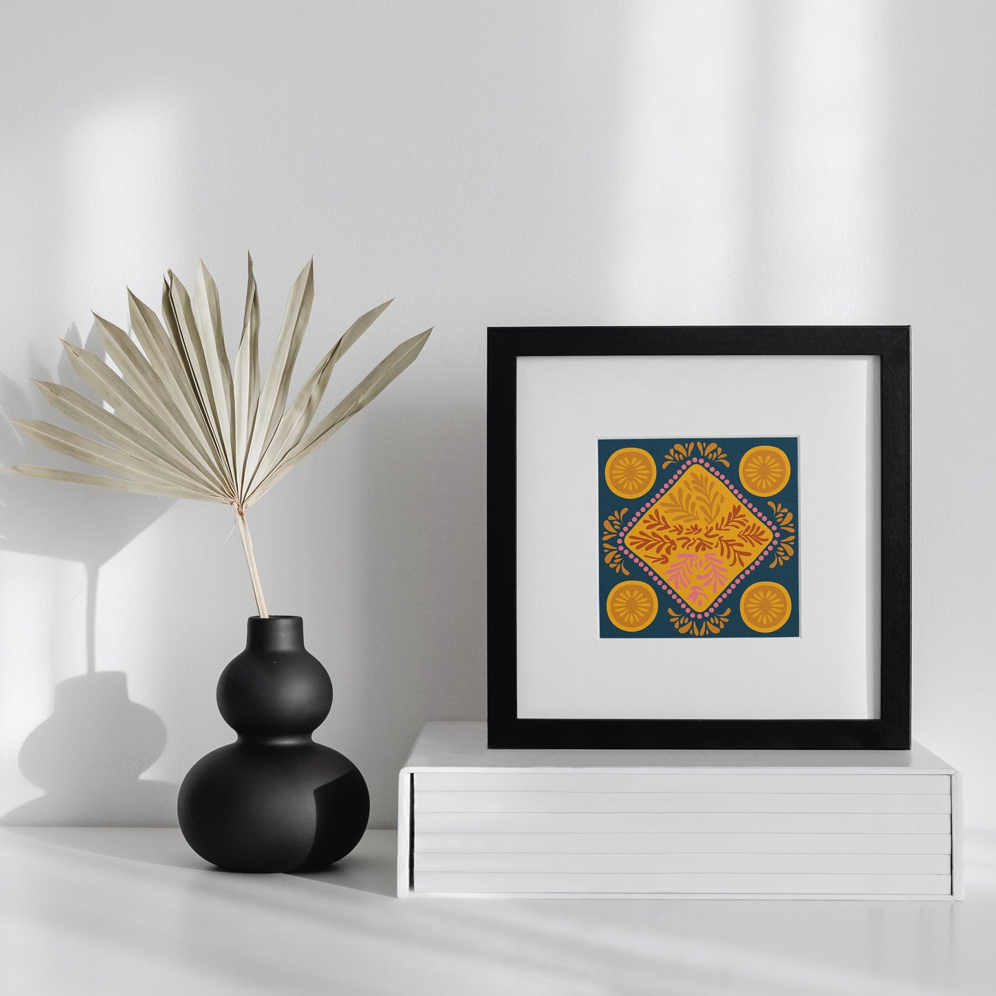Framed artwork on a white surface with a black vase and dried plant.
