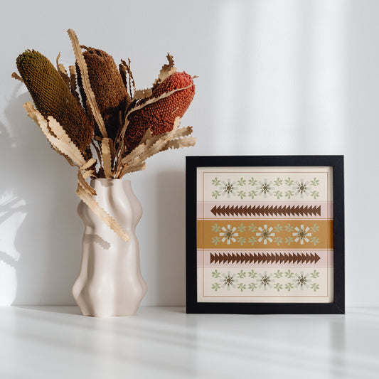 Decorative vase with dried plants next to a framed artwork on a white surface.