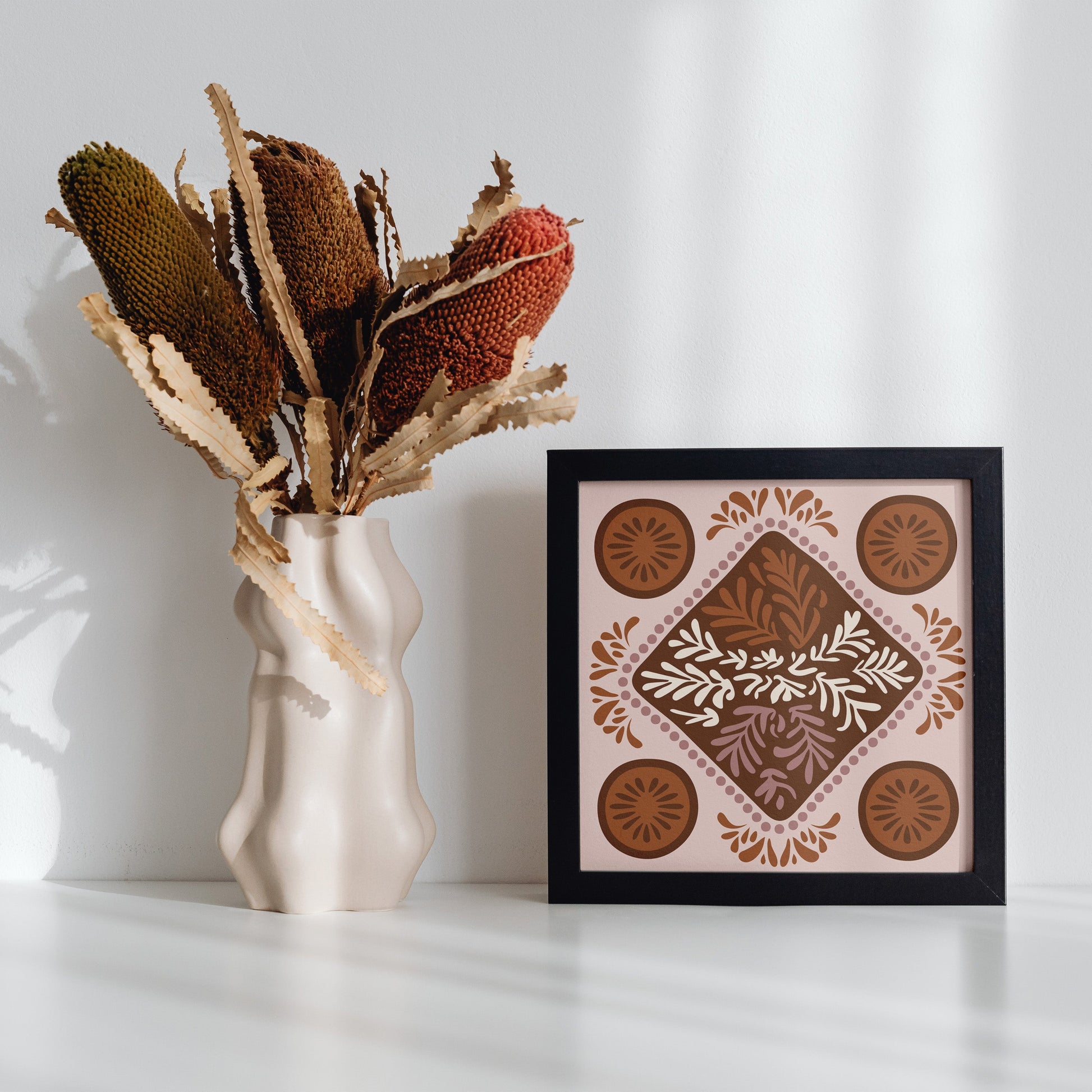 Decorative vase with dried plants next to a framed artwork on a white surface.