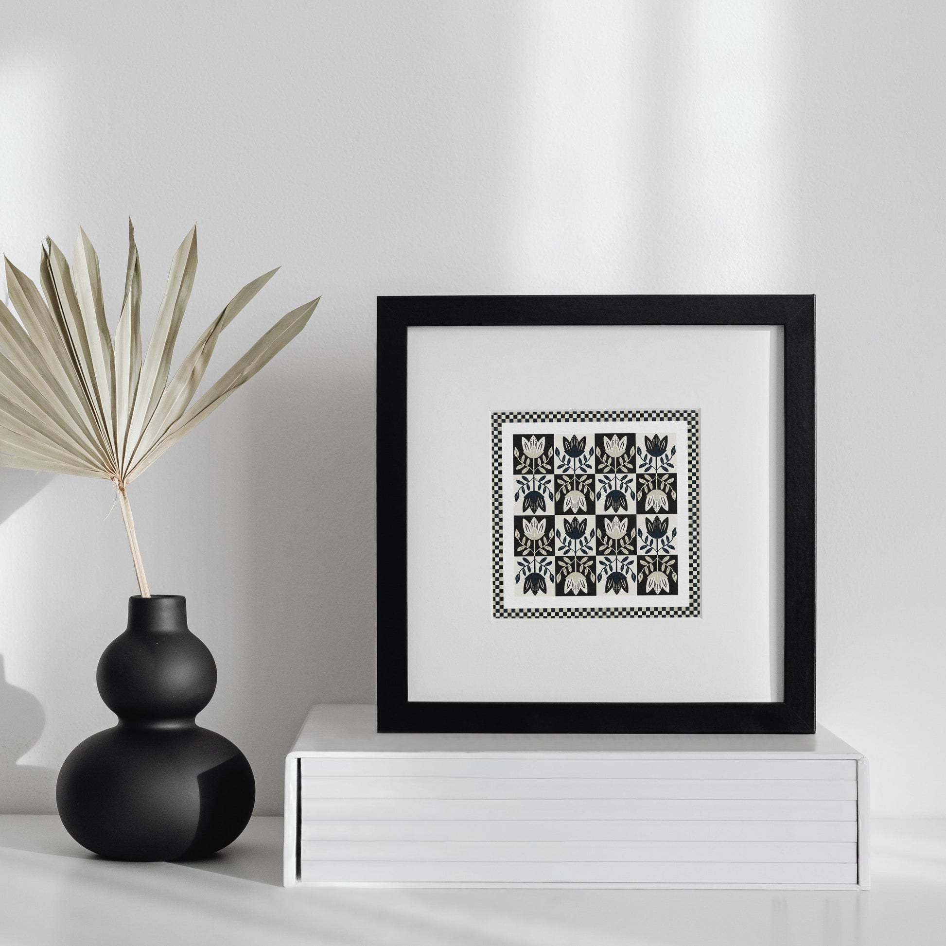 Framed artwork on a white surface with a black vase and dried plant.