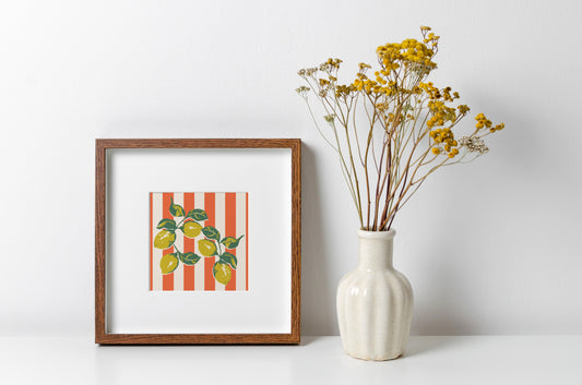 Framed artwork with striped pattern and lemons next to a vase with yellow flowers on a white surface.
