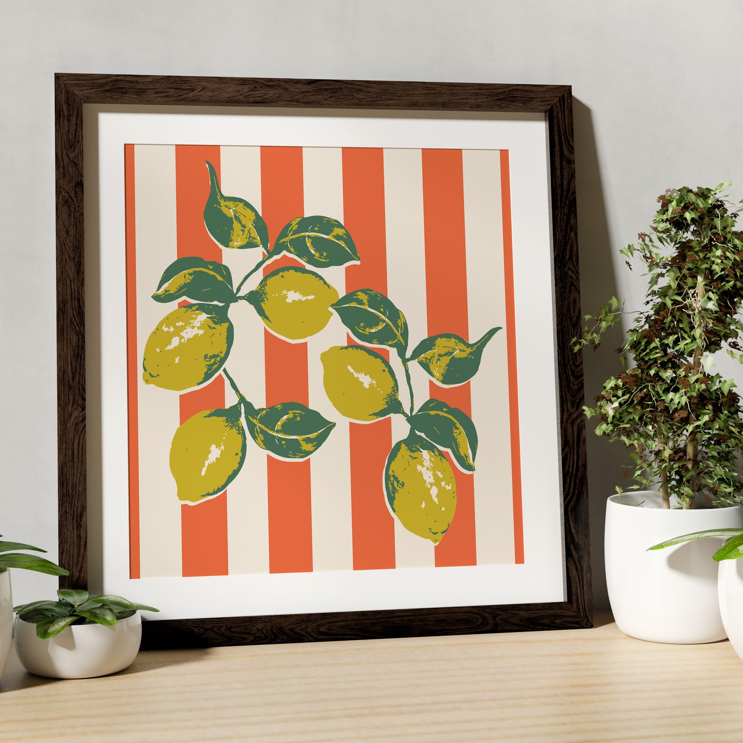 Framed artwork with lemon design on a striped background, placed on a wooden surface with plants.