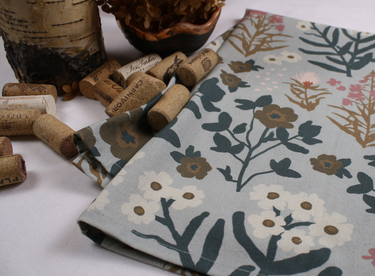 A folded fabric napkin or cloth with a botanical floral print in muted blue-gray, cream, brown, and pink, styled with wine corks and dried foliage on a white surface.