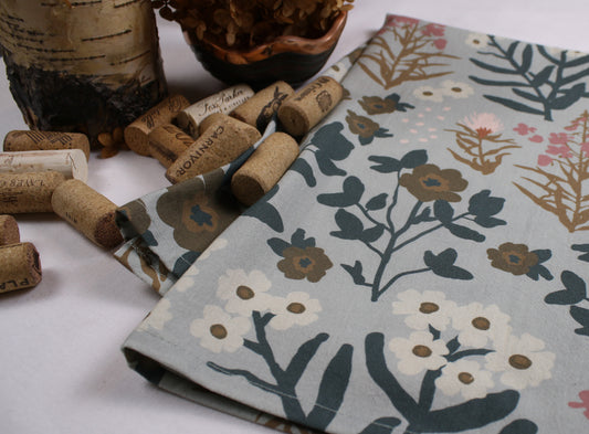 A folded fabric napkin or cloth with a botanical floral print in muted blue-gray, cream, brown, and pink, styled with wine corks and dried foliage on a white surface.